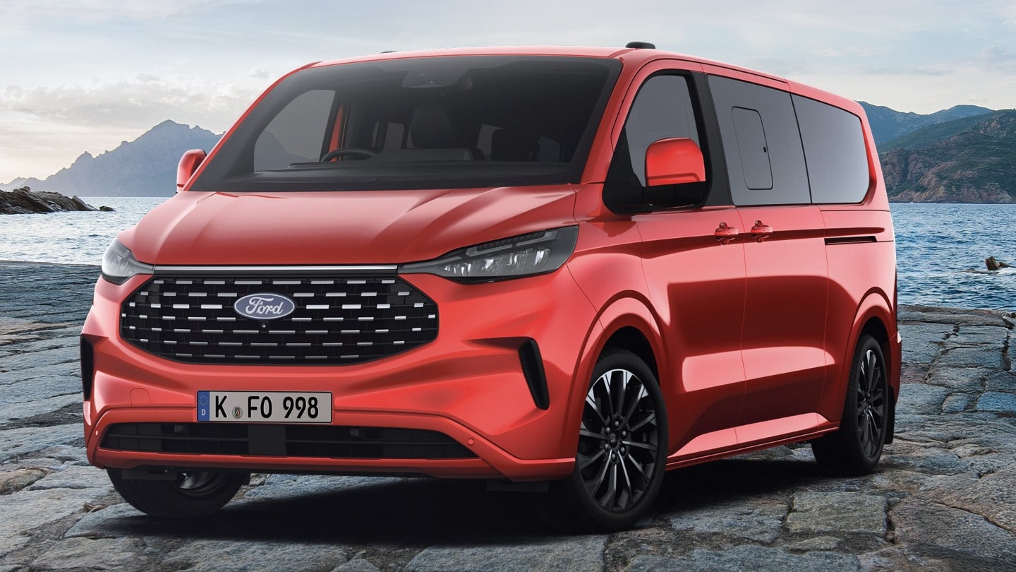 On peut voir un Ford Tourneo Custom en rouge, avec de l’eau et des sommets en arrière-plan.