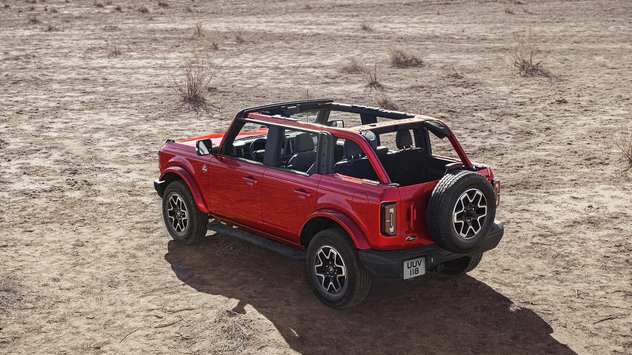 Ford Bronco en rouge. En vue 3/4 arrière avec le toit retiré, debout sur une surface sablonneuse