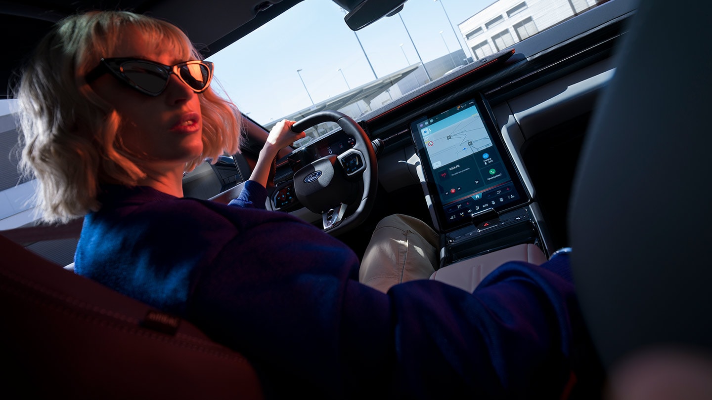 Une femme conduit la Ford Capri® entièrement électrique. On voit le volant, le tableau de bord et le grand écran tactile Ford SYNC® Move.