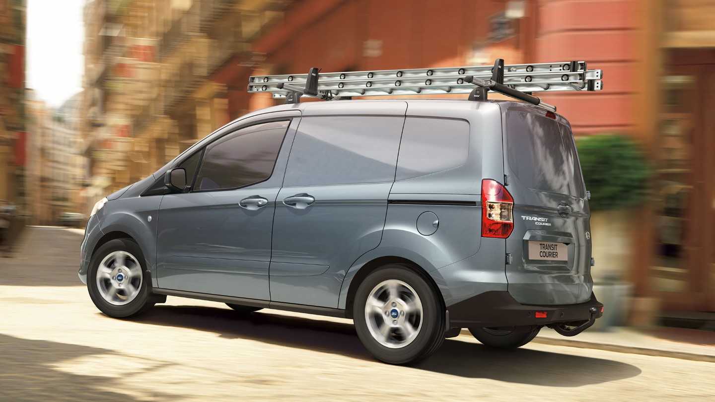 Ford Transit Courier, couleur bleu argenté, sur une route avec galerie de toit et échelle. Vue arrière aux trois quarts.