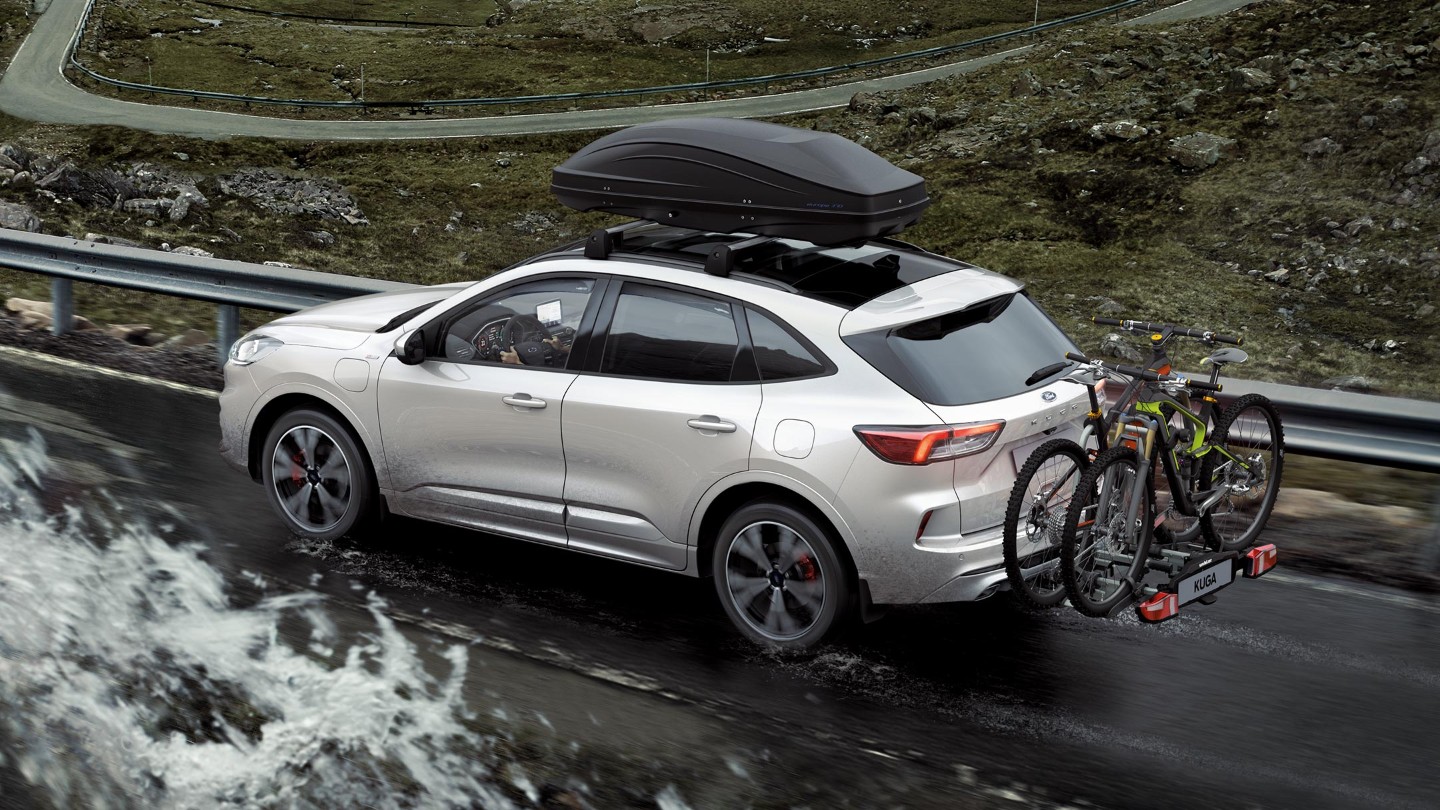 Ford Kuga, couleur argent, roulant sur une route de montagne avec un porte-vélos arrière et des vélos. Vue arrière aux trois quarts.