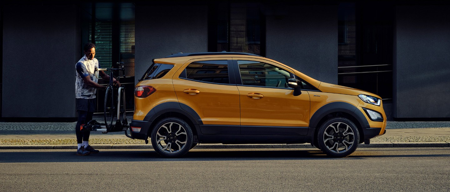 Vue latérale du Ford EcoSport couleur orange, parqué dans une rue en ville pendant qu’un homme fixe un vélo sur le porte-vélos arrière.